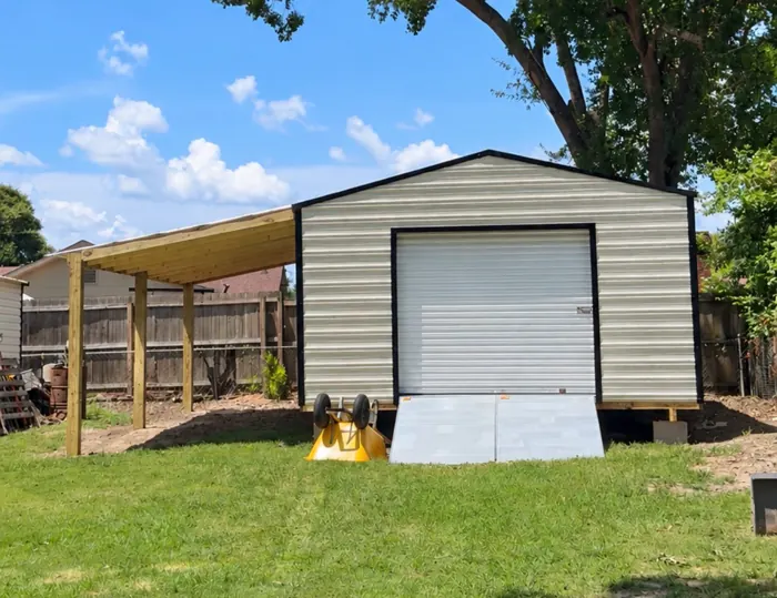 16x24 aluminum shed with a roll-up door, an aluminum ramp and a wood lean-to built on the side, under a large shade tree