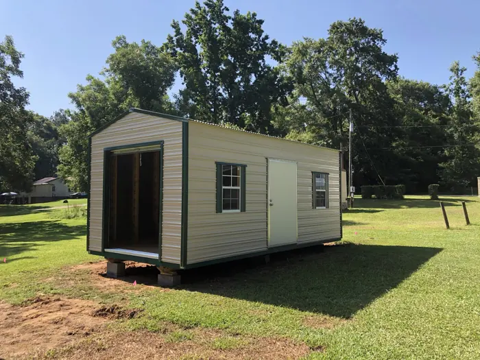 Cream aluminum shed with green trim and a roll-up door, leveled on concrete blocks