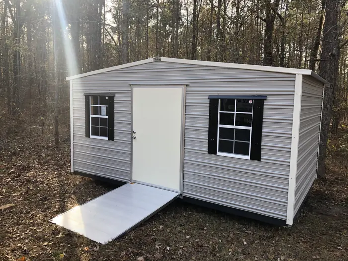 Grey aluminum shed with cream trim, black shutters and an aluminum ramp in the woods