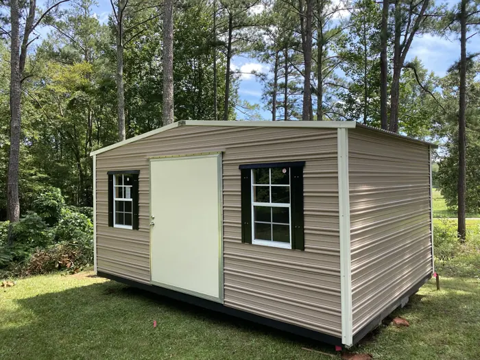 Tan aluminum shed with cream trim and black shutters among pine trees