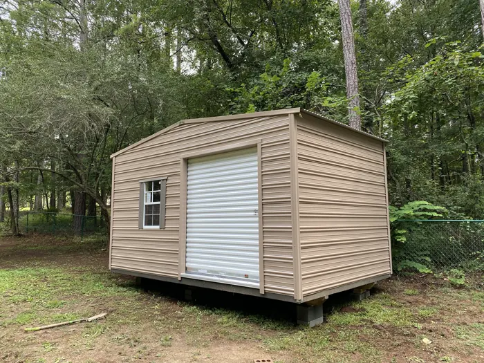 Tan aluminum shed with a roll-up door and one window, leveled on concrete blocks in a wooded yard