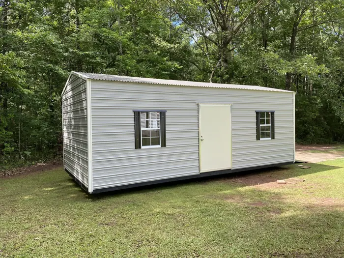 White aluminum shed with black shutters and a single door in a wooded yard