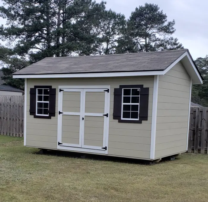 Beige wood shed with lap siding, dark brown shutters and double doors in a fenced yard
