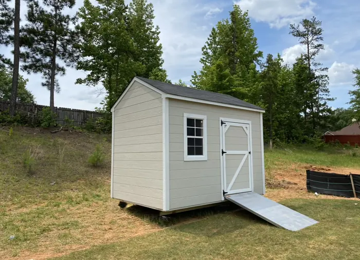 Beige wood shed with lap siding, a Z-braced door and an aluminum ramp on a sloped backyard
