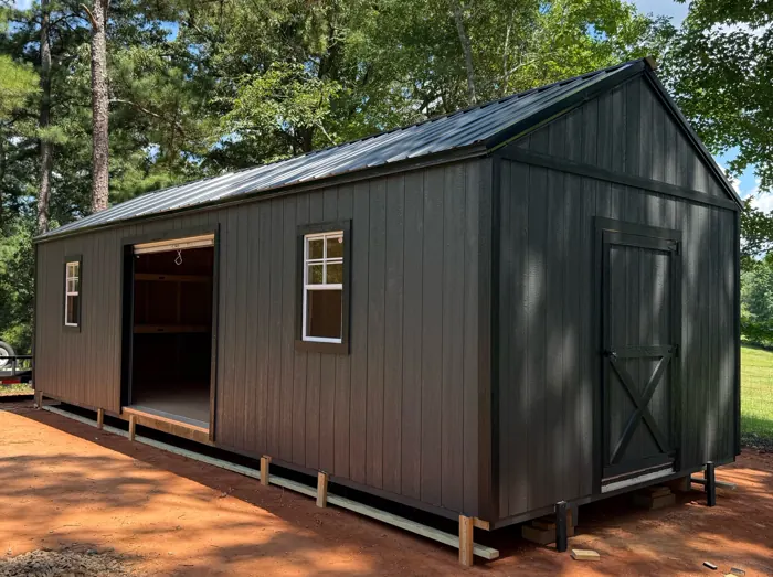 Long black wood shed with a black metal roof, a roll-up door and an X-braced end door, built on a cleared lot among pines