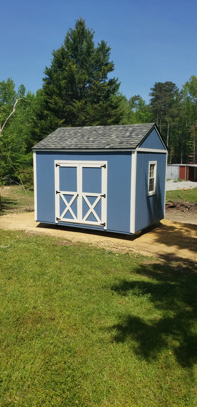 Blue wood shed with white X-braced double doors and a shingle roof on a cleared pad