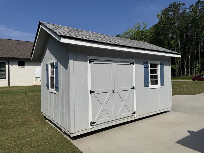 Light grey wood shed with blue window shutters and X-braced double doors beside a driveway