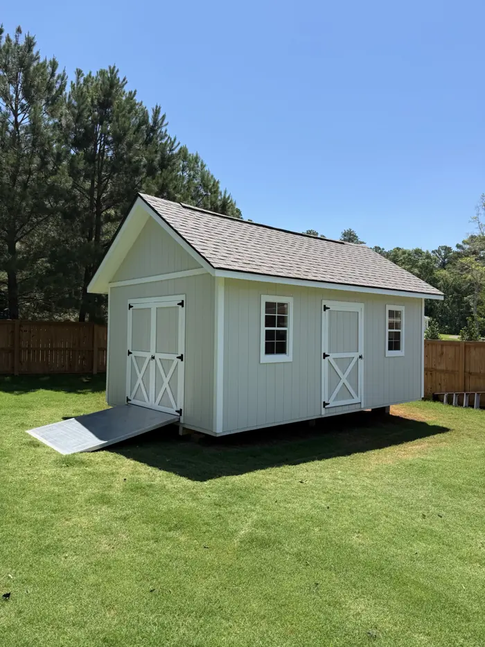 Light grey wood shed with double doors on the end, a second door on the side and an aluminum ramp in a fenced backyard