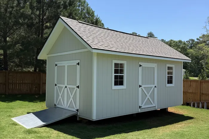 Grey wood shed with white trim, X-braced double doors on the end, a second door on the side, two windows and a shingle roof, with an aluminum ramp