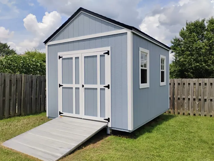 Light blue wood shed with double doors on the gable end and an aluminum ramp in a fenced yard