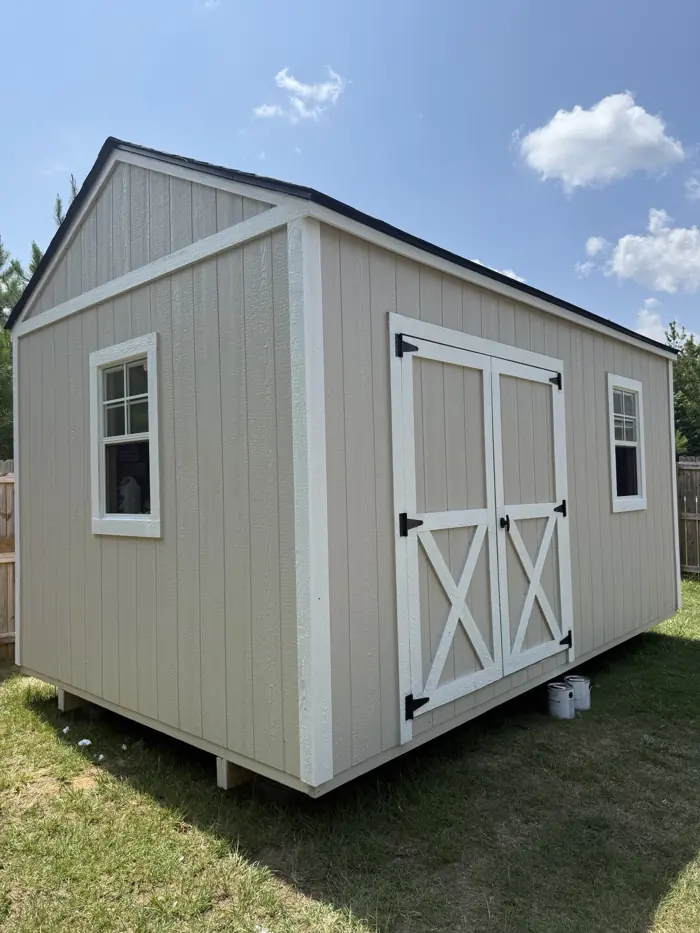 Tan wood shed with white X-braced double doors and white trim in a backyard