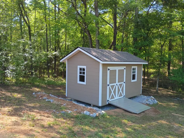 Tan wood shed with white trim, X-braced double doors, two windows and a shingle roof, built on a gravel pad with an aluminum ramp in a wooded backyard