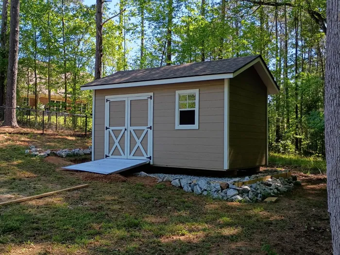 Side view of a tan wood shed with white trim, double doors, a ramp and a stone-edged gravel pad