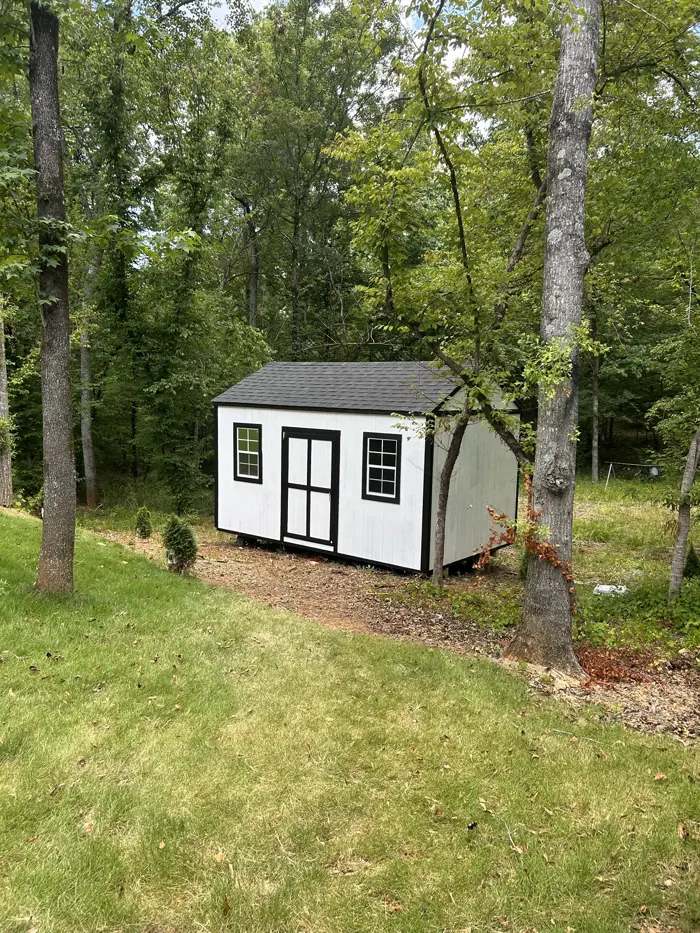 White wood shed with black trim and a black shingle roof among trees